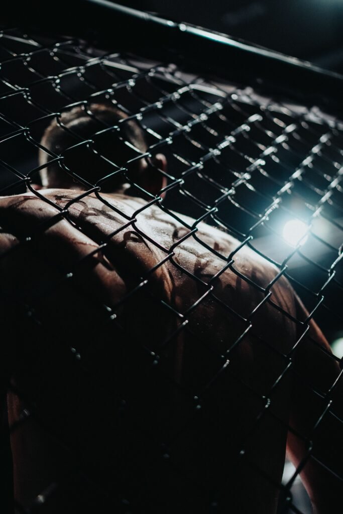 UFC 329: The Return of the Mac. Moody shot of a muscular MMA fighter seen through the cage at night, Vienna.